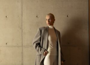 Mulher posa em frente a uma parede de concreto aparente, explorando tons e texturas. Ela veste um casaco de lã cinza sobre uma blusa de gola alta e calça off-white, criando contraste entre o tecido estruturado do casaco e a leveza do look justo.