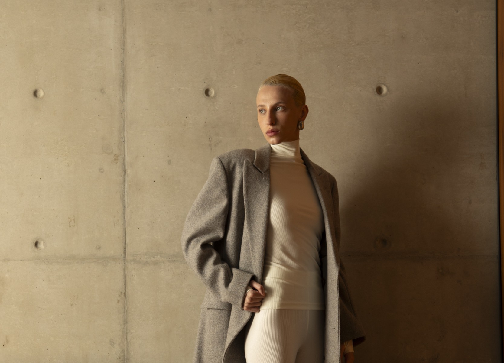 Mulher posa em frente a uma parede de concreto aparente, explorando tons e texturas. Ela veste um casaco de lã cinza sobre uma blusa de gola alta e calça off-white, criando contraste entre o tecido estruturado do casaco e a leveza do look justo.