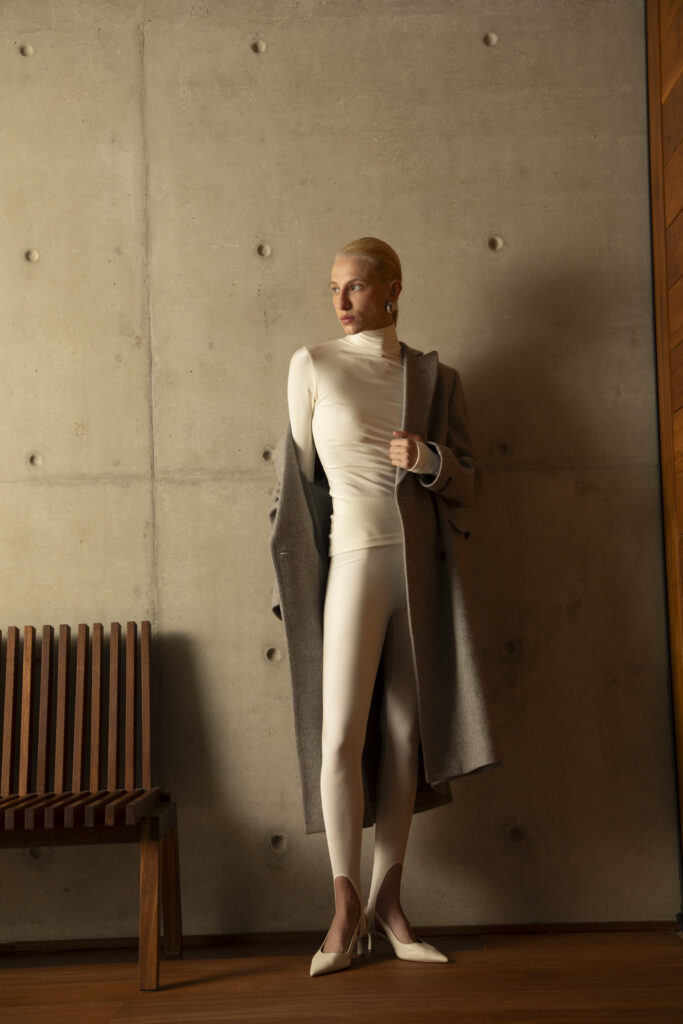 Modelo veste look monocromático em tons neutros, com conjunto off-white de cacharrel e calça canelada, sobreposto por casaco de alfaiataria cinza.