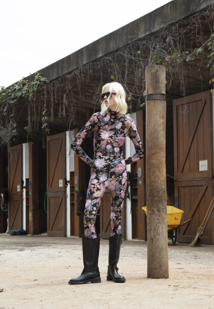Modelo usando macacão em lycra estampado floral de alta compressão, modelagem ajustada ao corpo, manga longa e efeito modelador, combinado com botas de cano alto, em ambiente externo rústico.