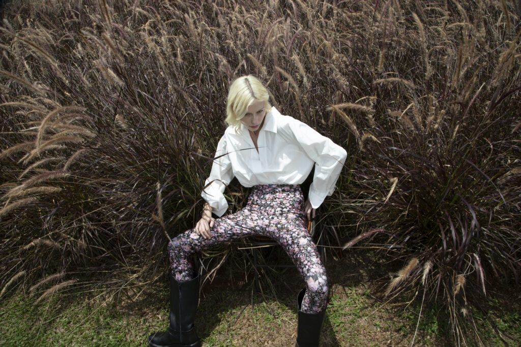 Mulher de cabelo loiro curto sentada em meio a vegetação ornamental, usando camisa branca, legging floral e botas pretas, em ensaio de moda ao ar livre.