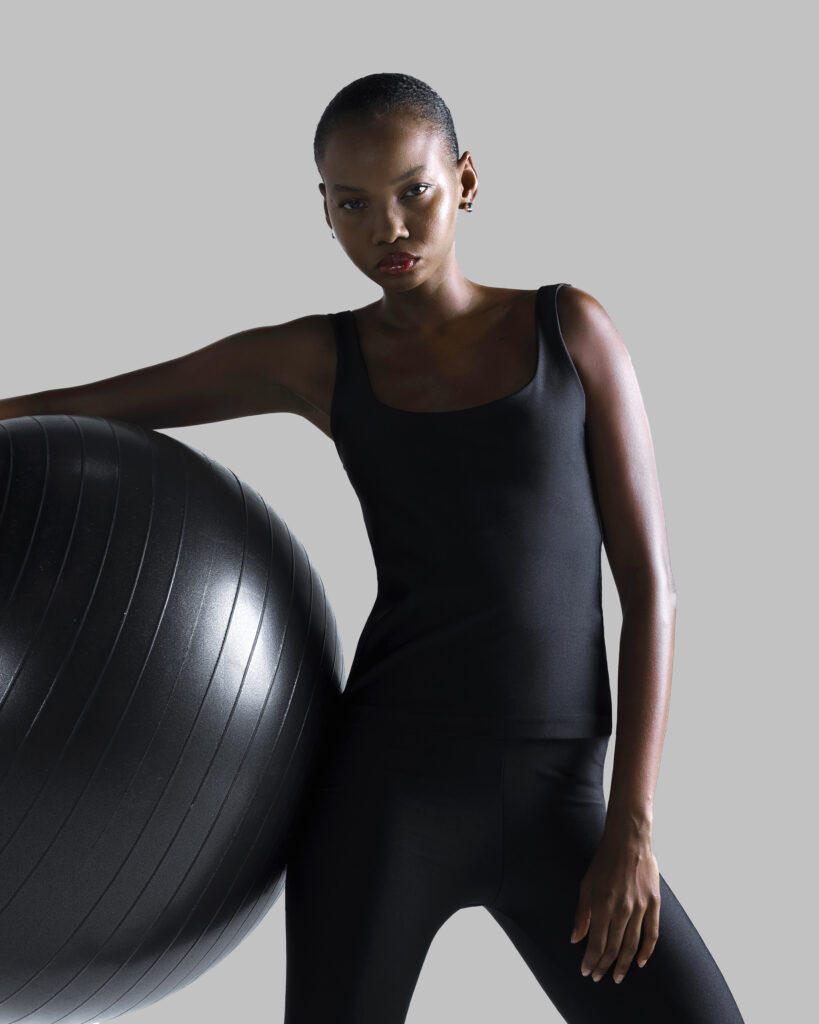 Mulher negra usando legging preta de lycra e regata preta ajustada, apoiada em uma bola de exercícios preta, em fundo neutro claro.