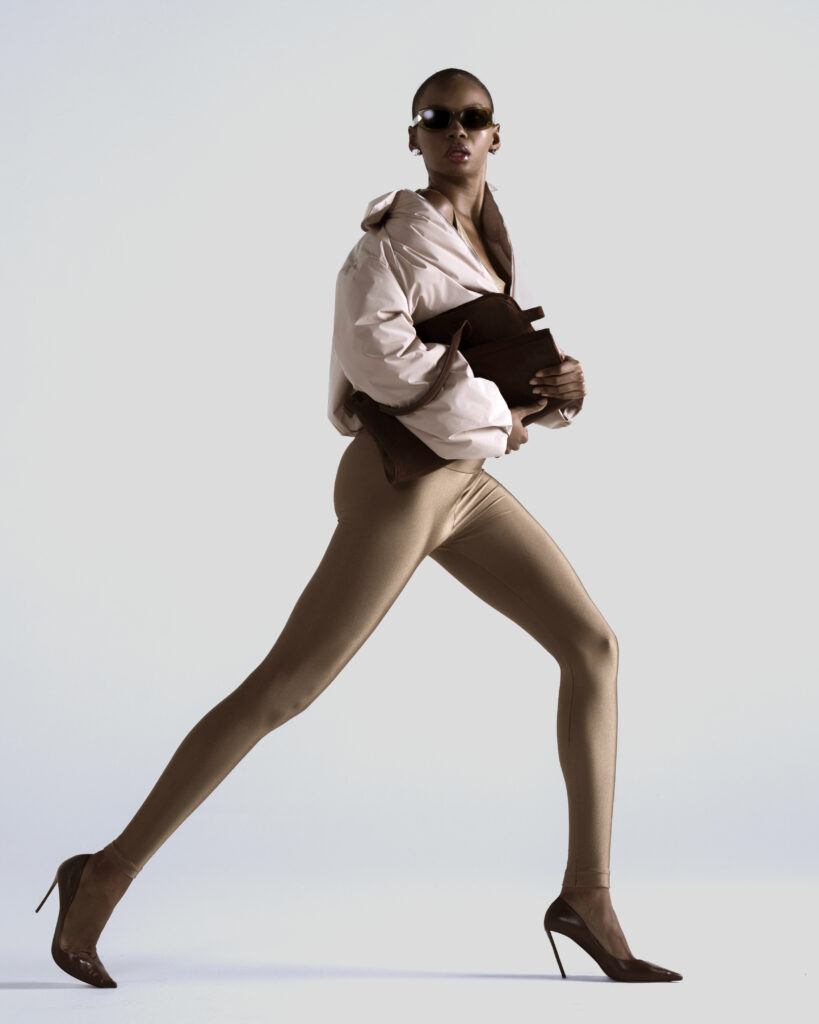 Mulher negra em pose de caminhada estilizada usando óculos escuros, jaqueta bege, legging de lycra nude, salto alto marrom e segurando uma bolsa marrom, em fundo neutro claro.