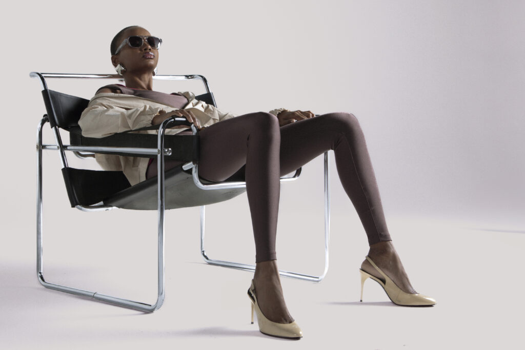 Mulher negra usando óculos escuros, jaqueta bege e legging marrom de lycra, sentada de forma relaxada em uma cadeira moderna de metal, usando salto alto bege, em fundo neutro claro.