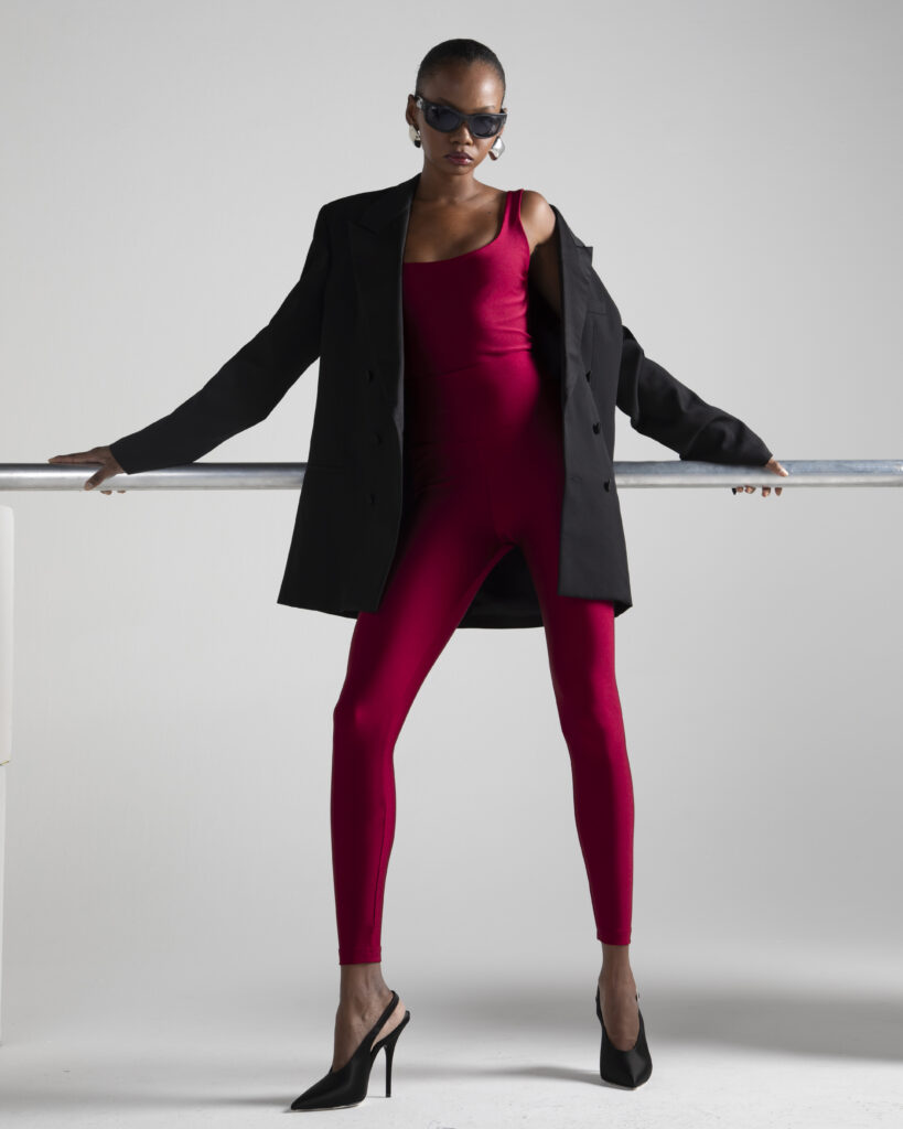 Mulher negra usando óculos escuros, blazer preto e legging vermelha de lycra, apoiada em uma barra metálica, com salto alto preto, em fundo neutro claro.
