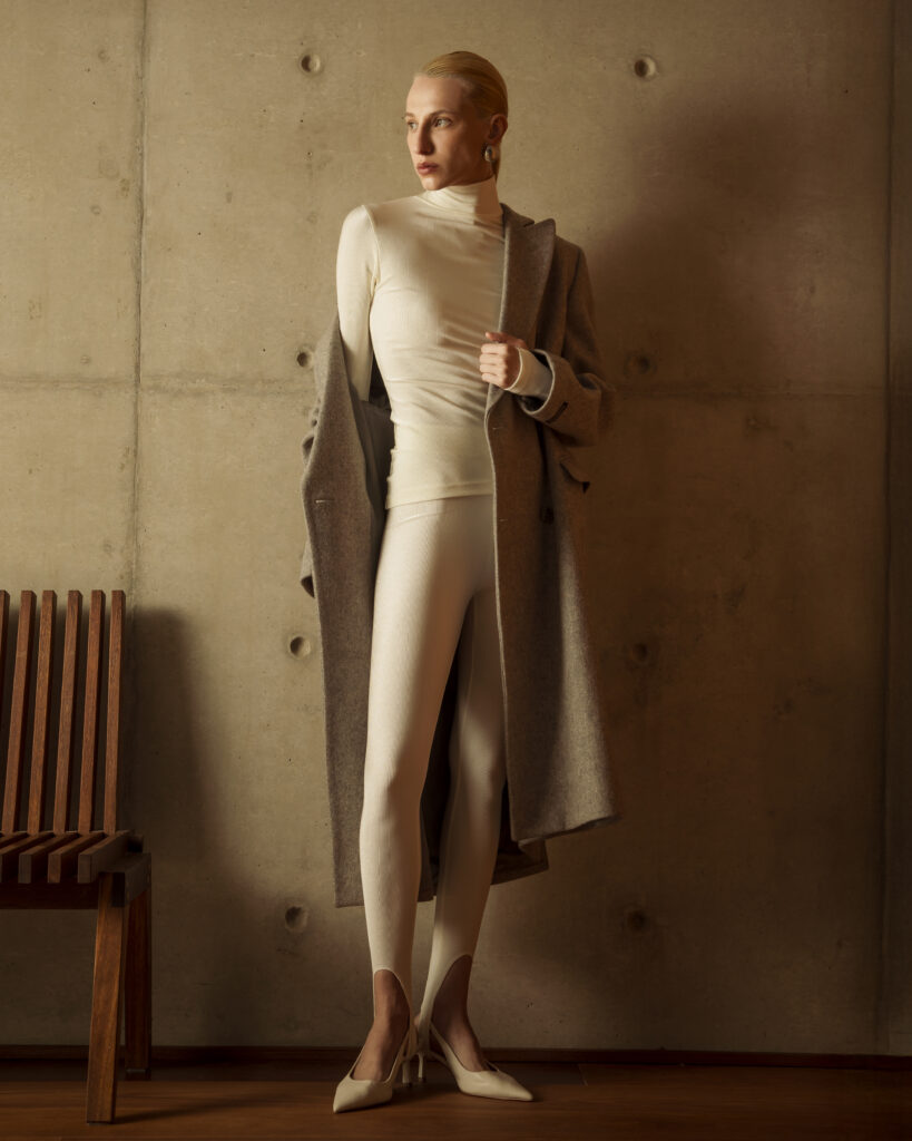 Mulher veste conjunto off-white com gola alta, casaco longo bege e sapatos claros em ambiente interno.