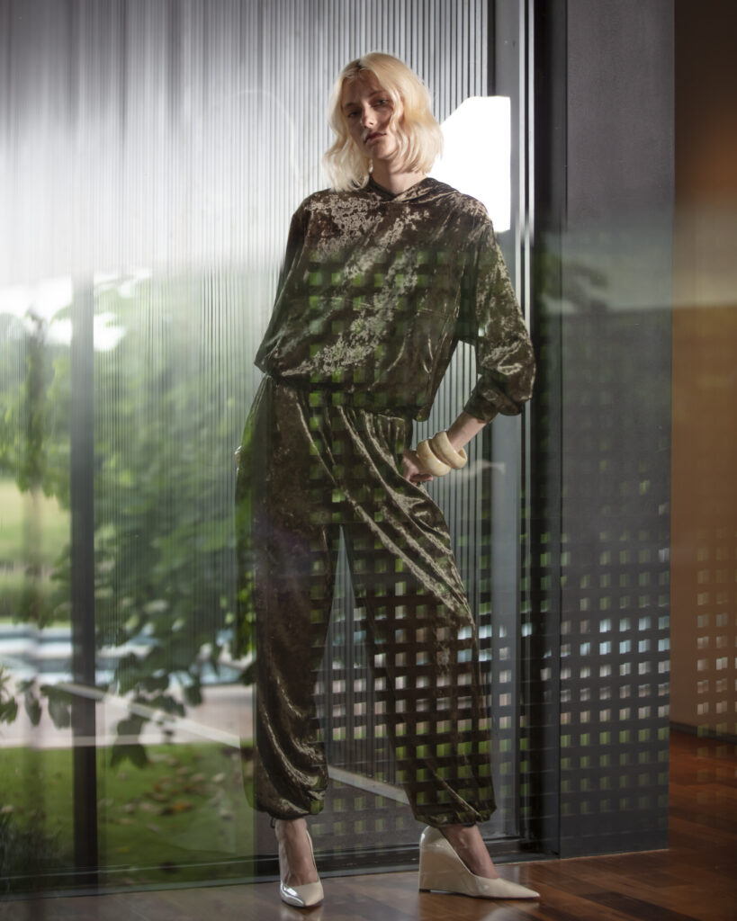 Mulher loira usando conjunto marrom de veludo com modelagem ampla e sofisticada, posando em ambiente contemporâneo com paredes de vidro texturizado, iluminação natural e acessórios em tons claros.