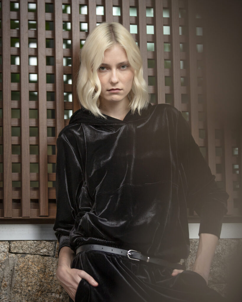 Close de mulher loira usando blusa preta de veludo com capuz, apoiada em estrutura de madeira com expressão séria e iluminação suave.