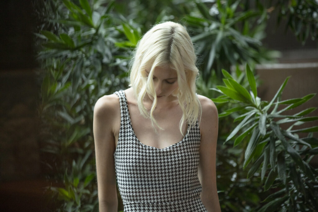 Modelo em plano aproximado, em ambiente externo com vegetação densa ao fundo. Ela está com a cabeça levemente inclinada para baixo, criando uma composição mais introspectiva. Veste uma peça única ajustada ao corpo, de alças finas e decote arredondado, com estampa clássica em padronagem Pied Poule (preto e branco), que traz sofisticação e um visual atemporal. O enquadramento destaca o caimento da peça na parte superior do corpo e a riqueza da padronagem em detalhe.