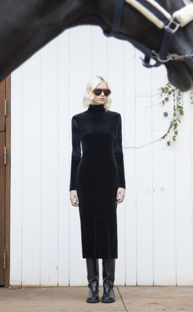 Modelo usa vestido preto de veludo com gola alta e mangas longas, combinado com botas e óculos escuros.