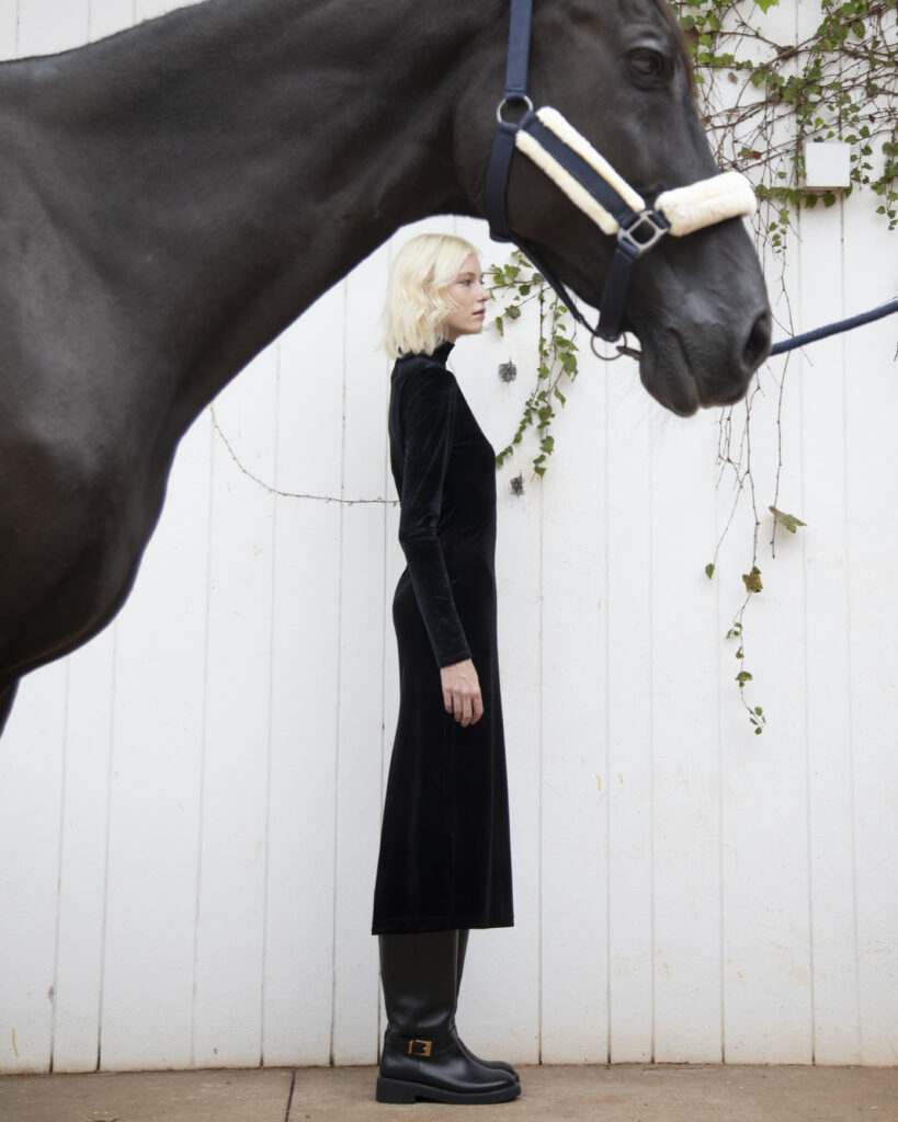 Mulher com vestido preto de veludo e botas posa de perfil ao lado de cavalo diante de parede branca.
