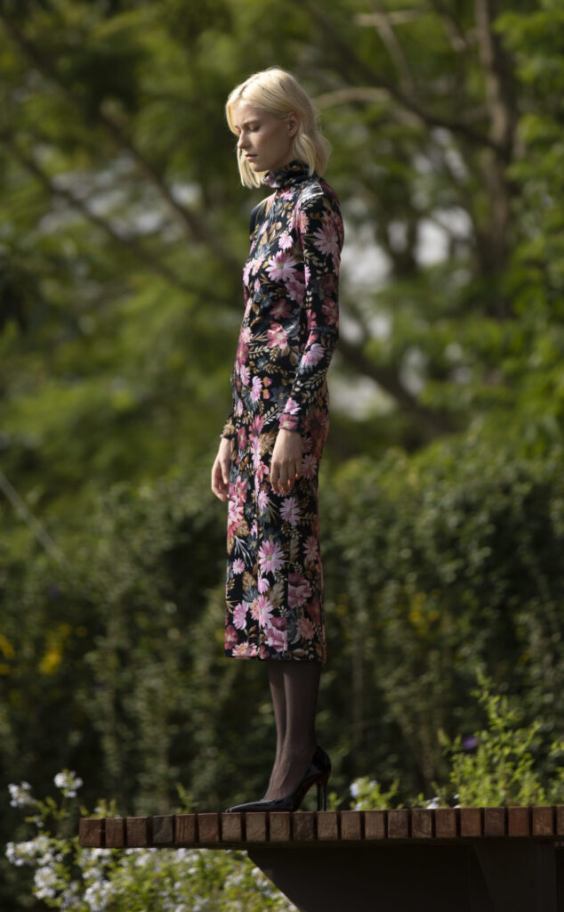 Mulher loira usando vestido floral de veludo com estampa em tons rosados, posando ao ar livre em jardim com árvores ao fundo.