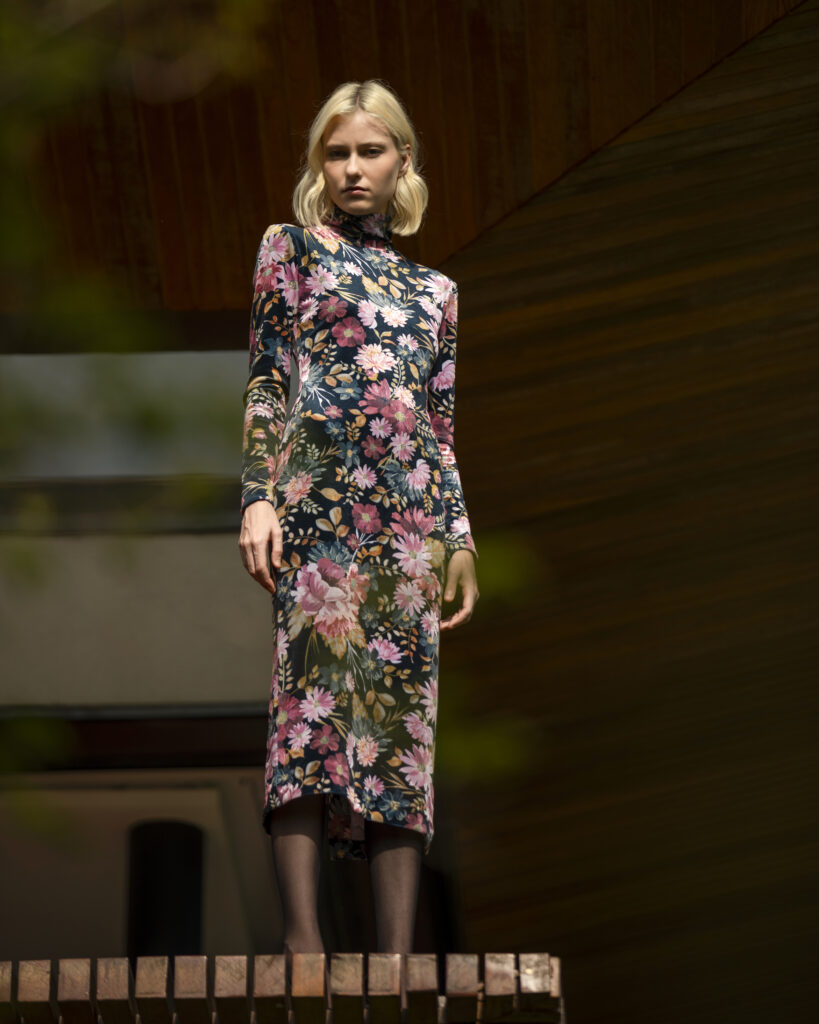 Mulher loira usando vestido midi de veludo floral com gola alta e mangas longas, em pé sobre estrutura de madeira diante de arquitetura contemporânea em tons escuros.