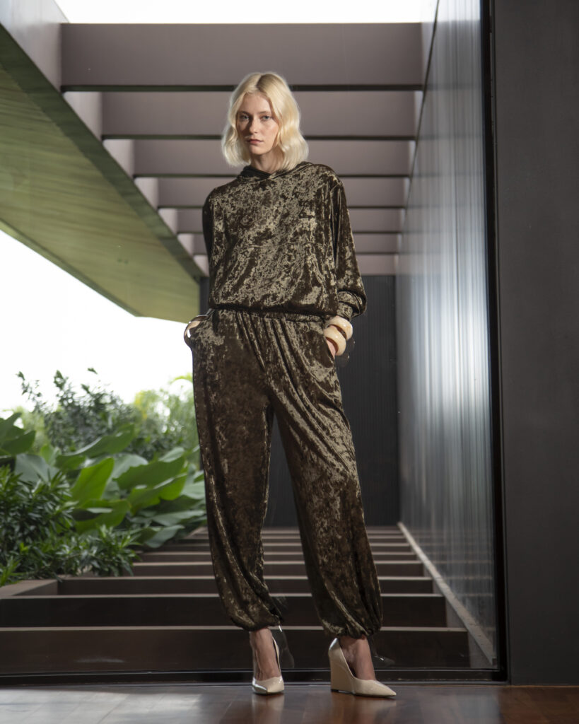 Modelo usa conjunto de veludo em tom bronze e sapatos claros em ambiente de arquitetura contemporânea.