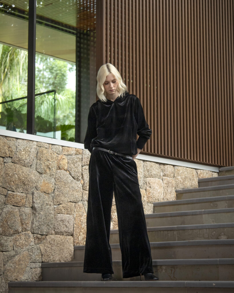 Mulher loira usando conjunto preto de veludo com modelagem ampla, posando sentada em escada de pedra ao lado de painel ripado de madeira.