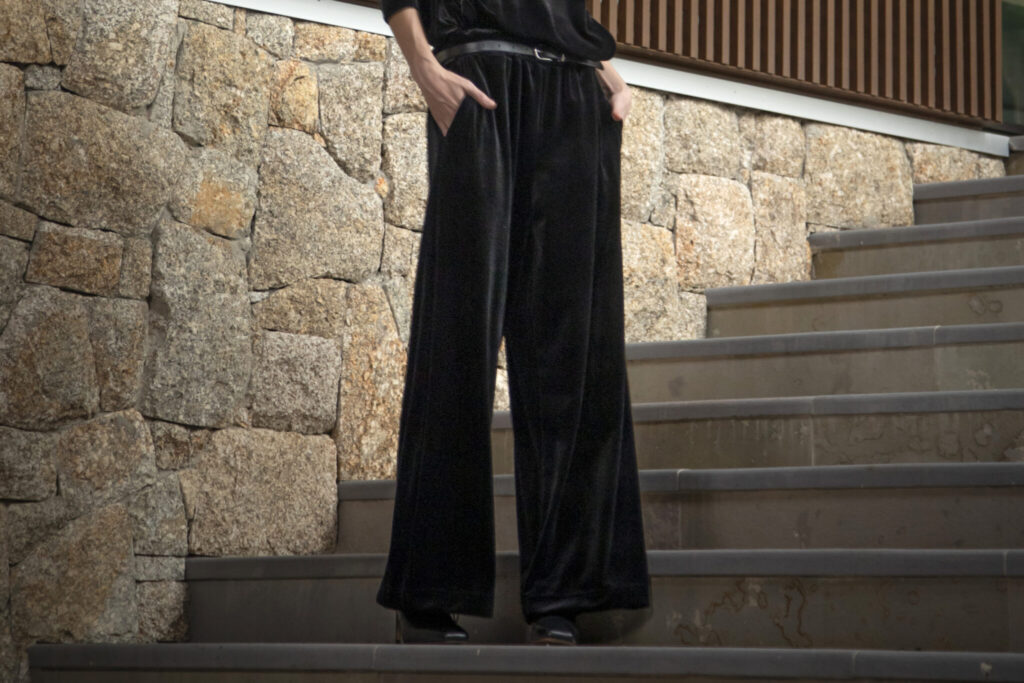 Calça pantalona de veludo preta em destaque sobre escadaria com parede de pedras e detalhes em madeira.