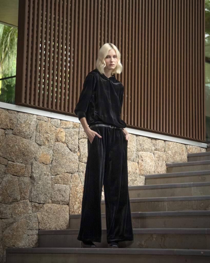 Mulher loira usando conjunto preto de veludo com blusa de mangas longas e calça pantalona de caimento fluido, posando em escada de pedra diante de arquitetura contemporânea com detalhes em madeira ripada.