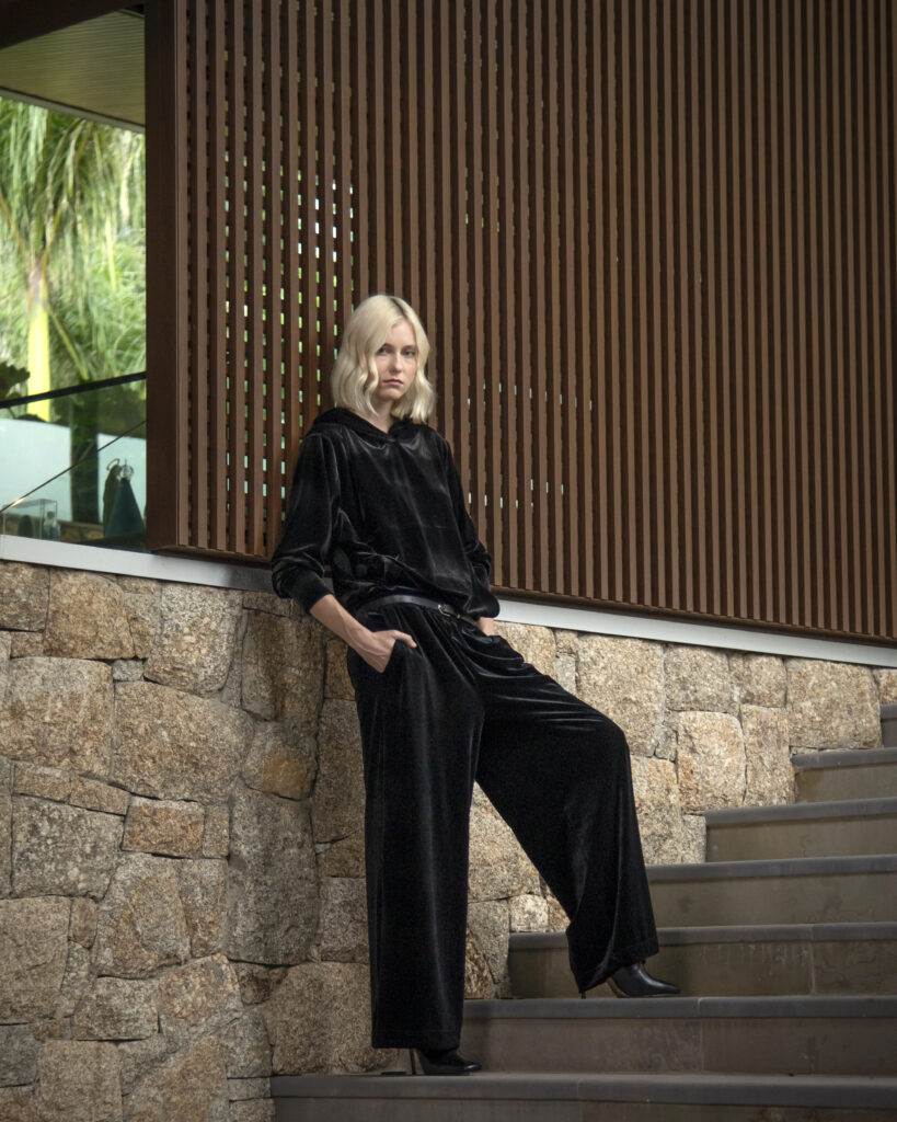 Mulher veste conjunto preto de veludo com modelagem ampla, posando em escadaria que destaca elegância, conforto e sofisticação.