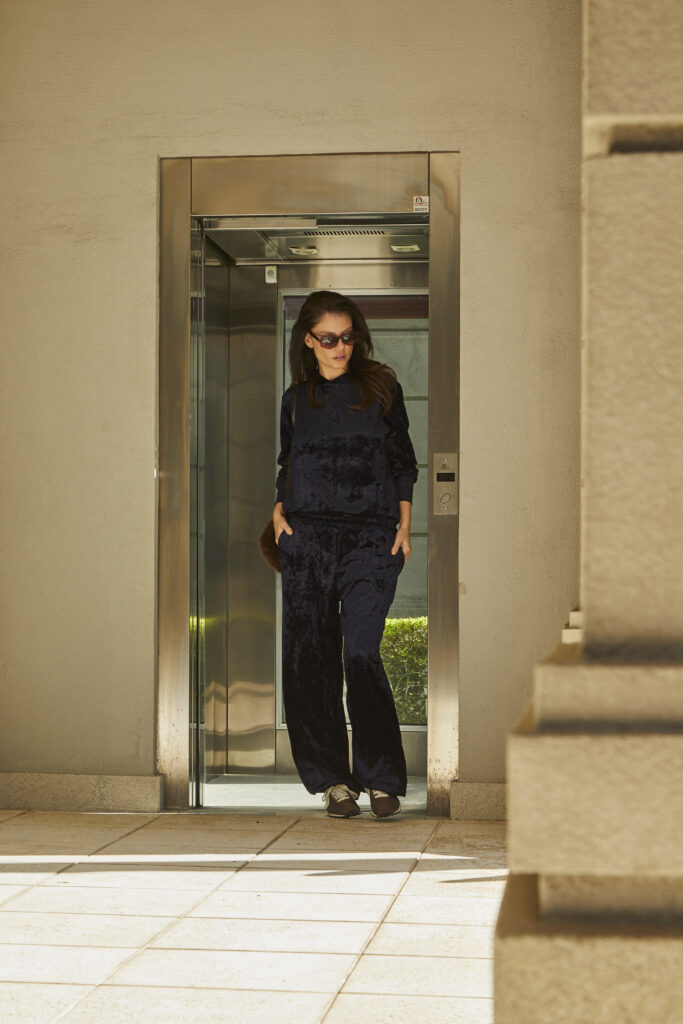 Mulher com conjunto azul-marinho de veludo, óculos escuros e modelagem ampla posa em ambiente externo elegante.