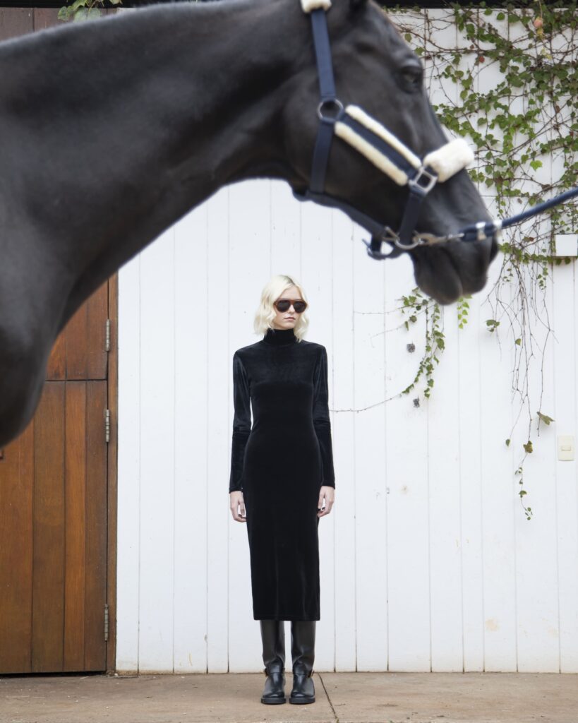 Mulher com vestido preto de veludo e óculos escuros posa diante de parede branca, com cavalo em primeiro plano.