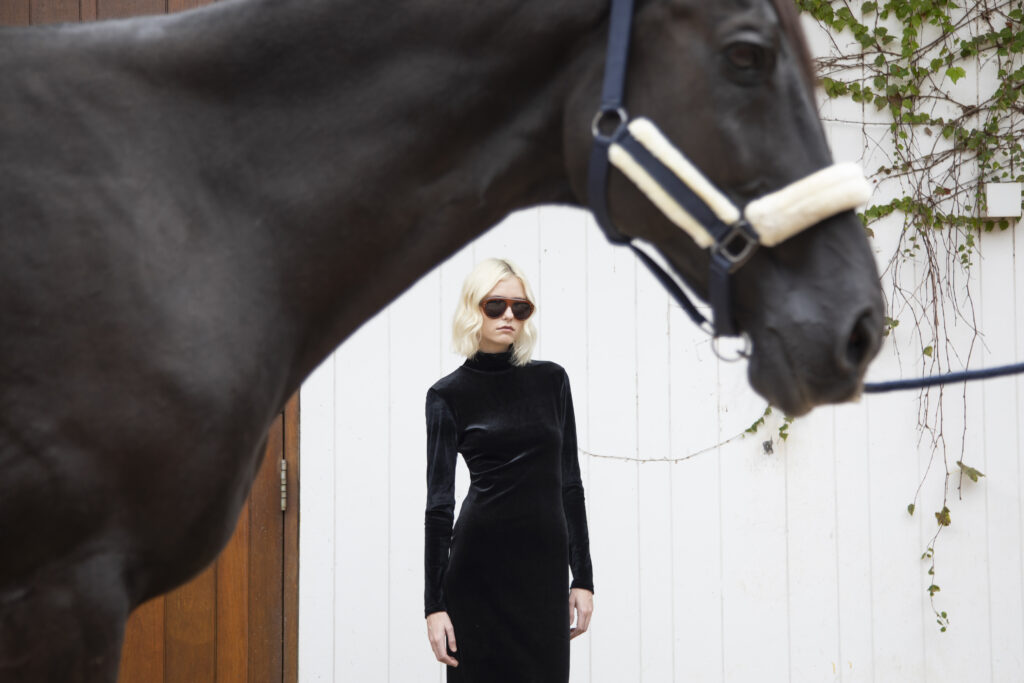 Mulher com vestido preto de veludo e óculos escuros posa ao fundo, enquanto um cavalo aparece em primeiro plano.