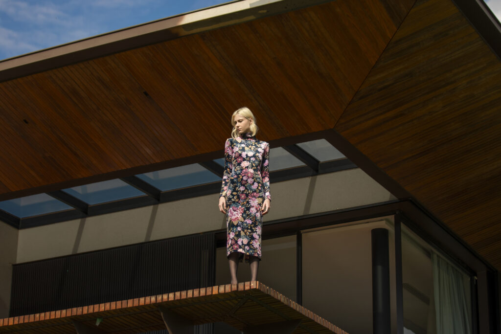 ulher com vestido de veludo floral posa em estrutura elevada diante de residência de arquitetura contemporânea.