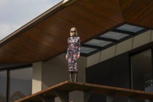 Mulher usando vestido de veludo estampado e óculos escuros posa em residência de arquitetura moderna.