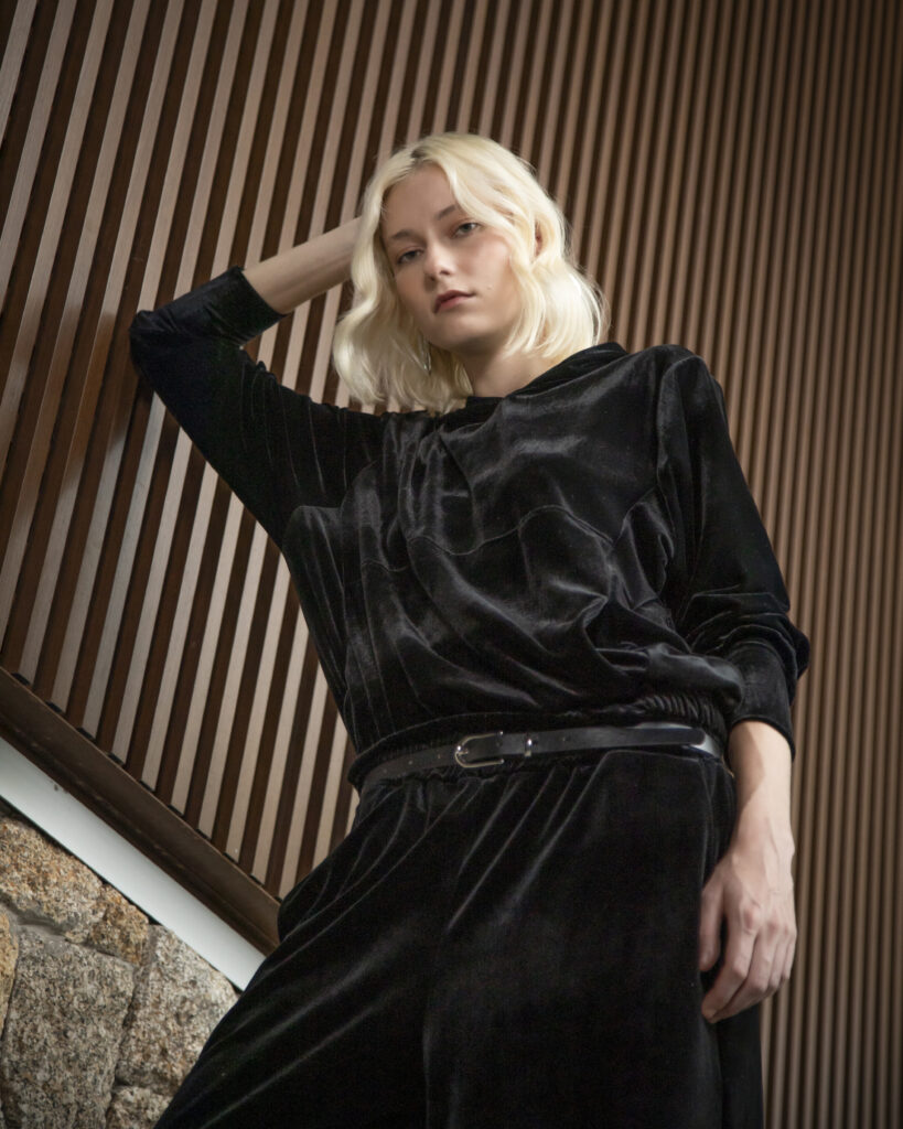 Mulher com vestido preto de veludo posa diante de painel de madeira, destacando a textura sofisticada e o caimento elegante da peça.