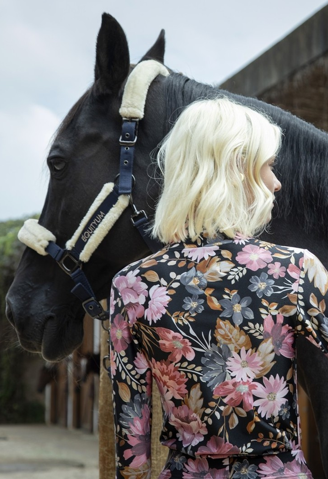 Modelo de costas com blusa floral ao lado de um cavalo preto, em ambiente externo de haras.