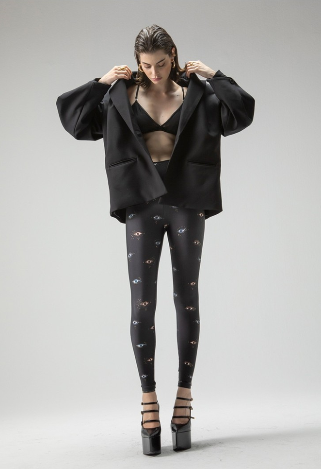 Modelo veste legging preta com estampa de olhos, blazer oversized e sandálias de salto em estúdio.