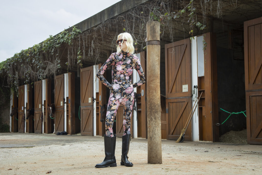 Mulher usa legging e blusa de gola alta com estampa floral, botas pretas e óculos em cenário de haras.
