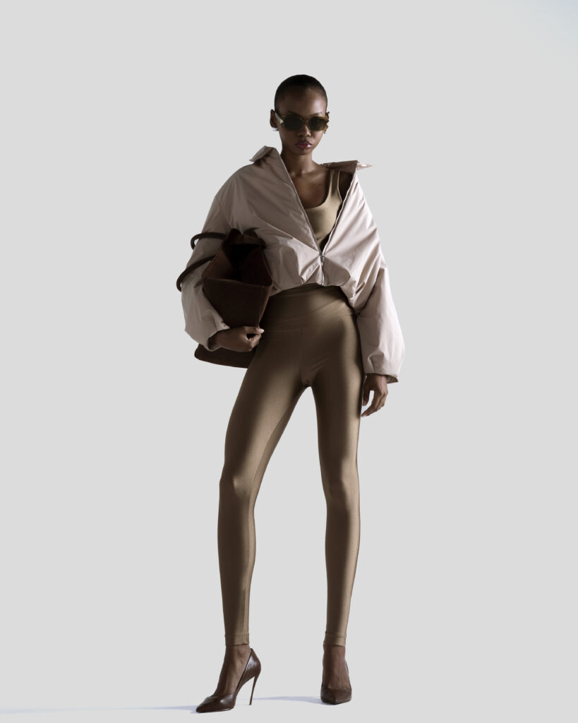 Mulher usa legging bege, top, jaqueta clara oversized, bolsa marrom e scarpin sobre fundo neutro.